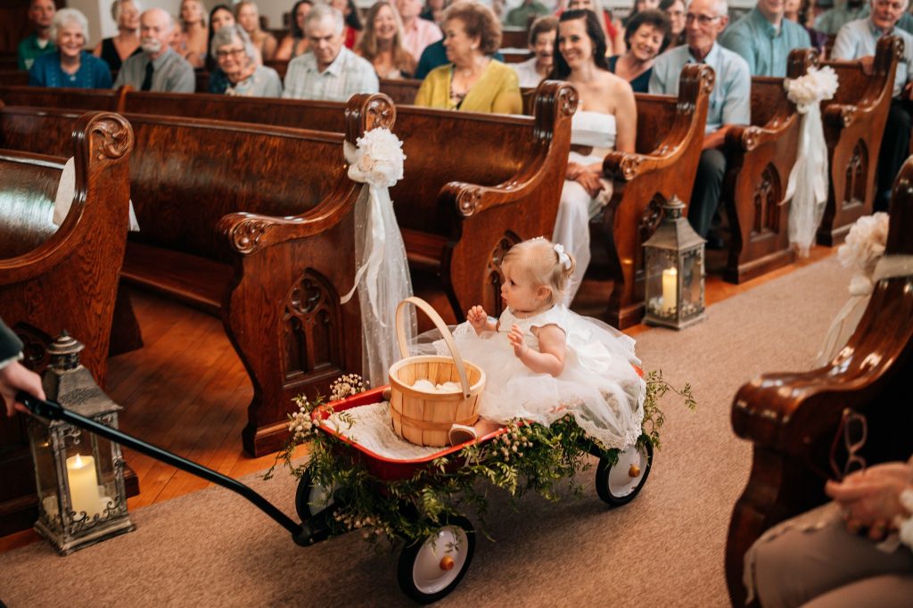 A flower girl riding in a wagon with a basket of flowers makes her way down the aisle of Devotions Wedding Chapel