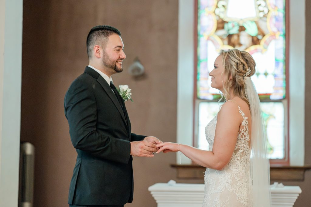 A bride and groom at the altar facing one another as the groom puts a ring on the brides' finger